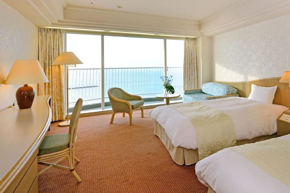 Habitación de hotel con dos camas, un escritorio, una silla, una mesita de noche y vista al mar.