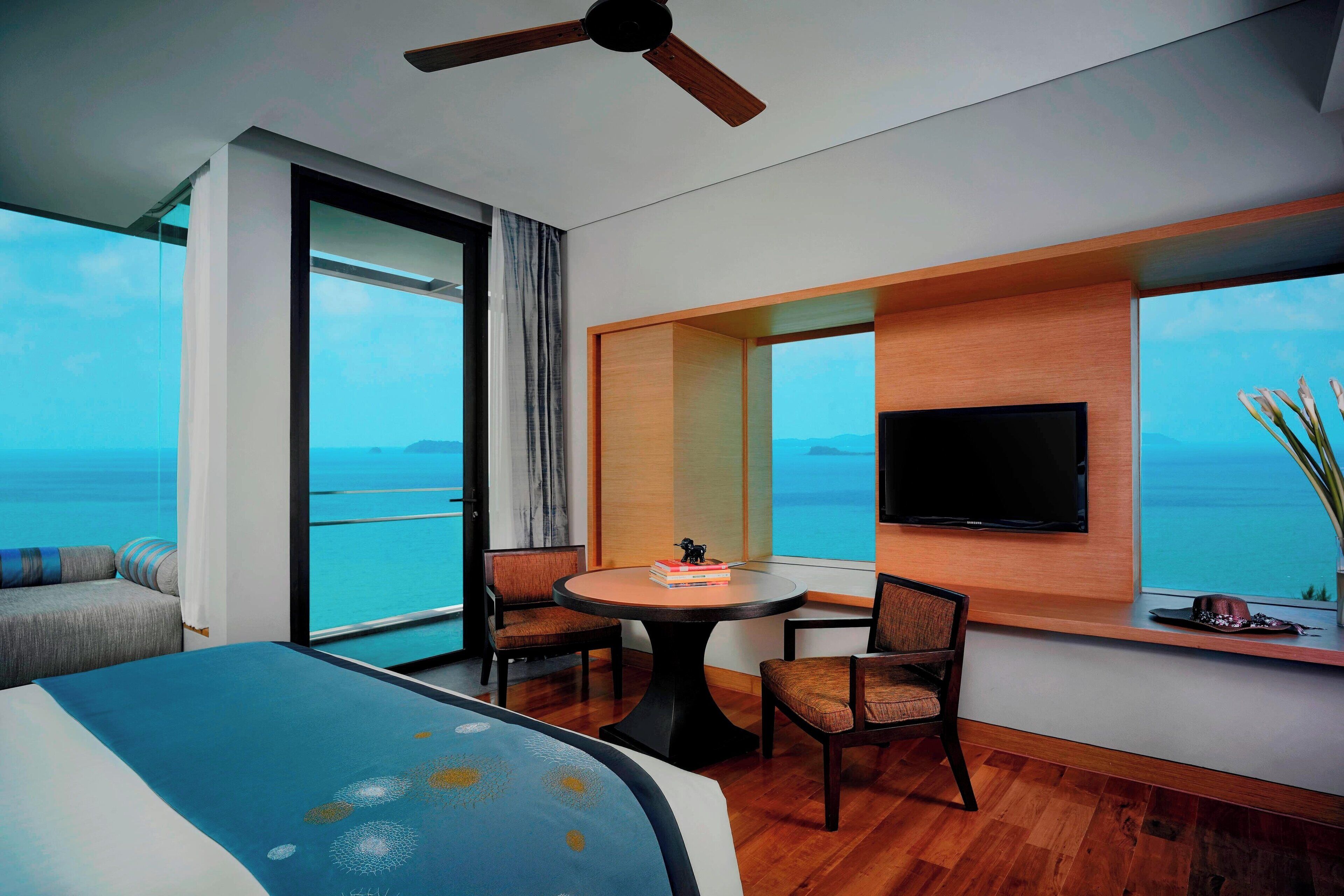 A modern hotel room with a large bed, a round table with chairs, a TV mounted on the wall, and a balcony with a view of the sea.