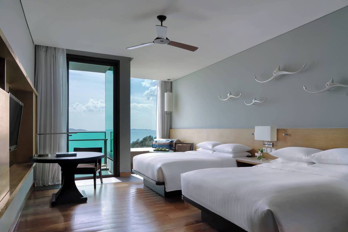A hotel room with two beds, a dining table, a ceiling fan, and a balcony with a view.
