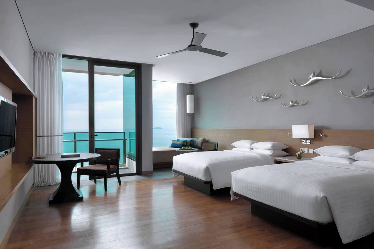 A hotel room with two beds, a dining table, a chair, a ceiling fan, and a large window with a view.