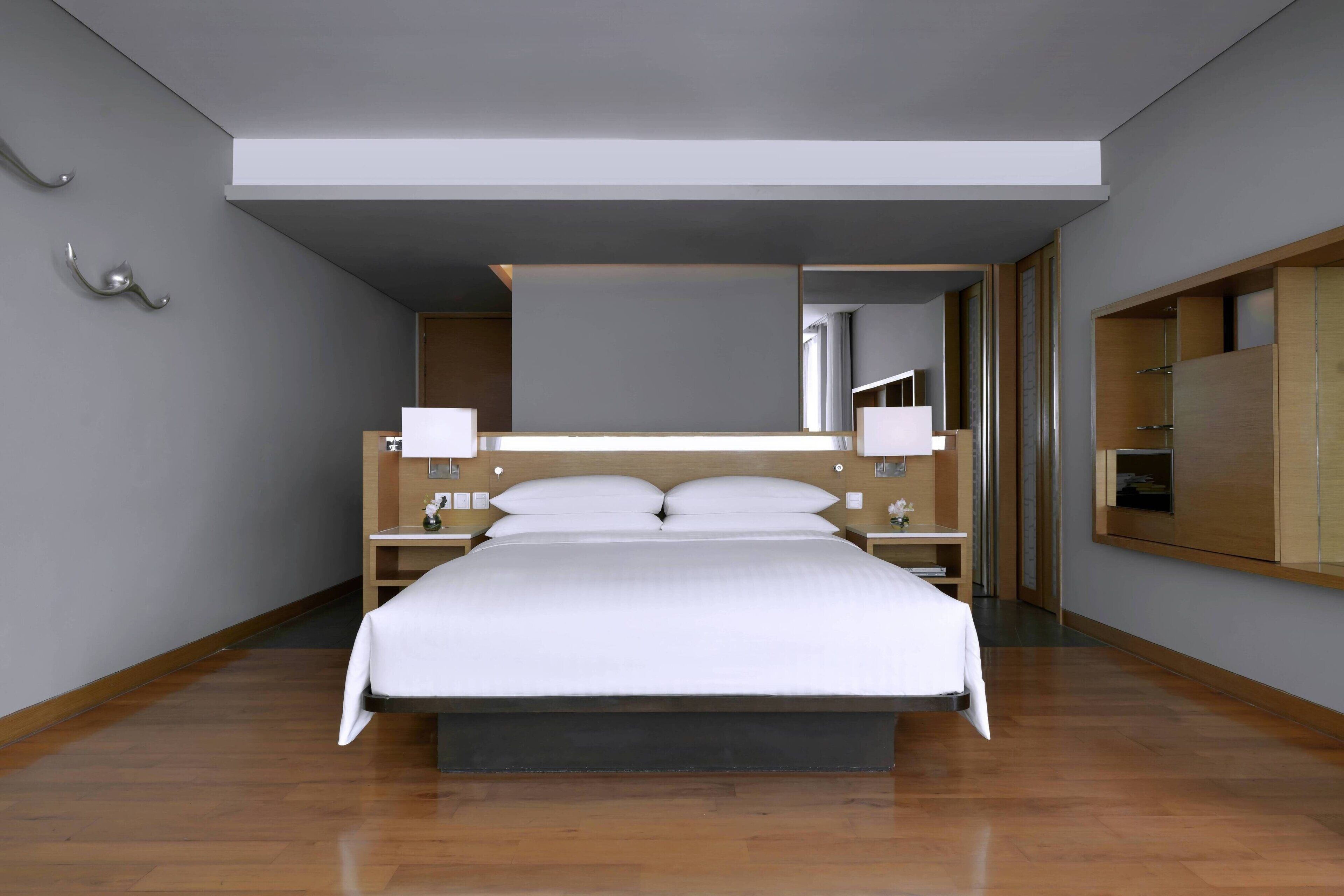 A modern hotel room with a large bed, wooden headboard, bedside tables with lamps, a built-in wardrobe, and a large mirror above the bed.
