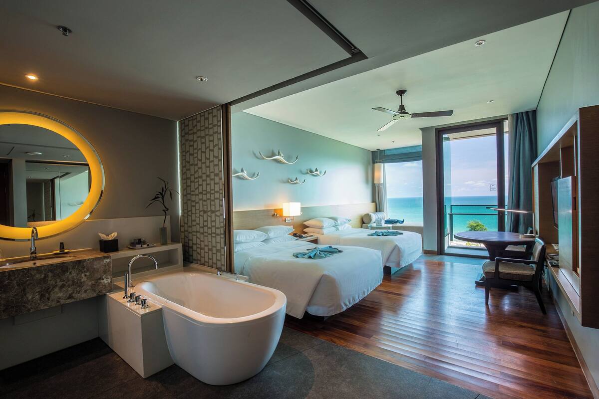A modern hotel room with a large bathtub, two beds, a balcony with a view, and a round mirror with a yellow light.