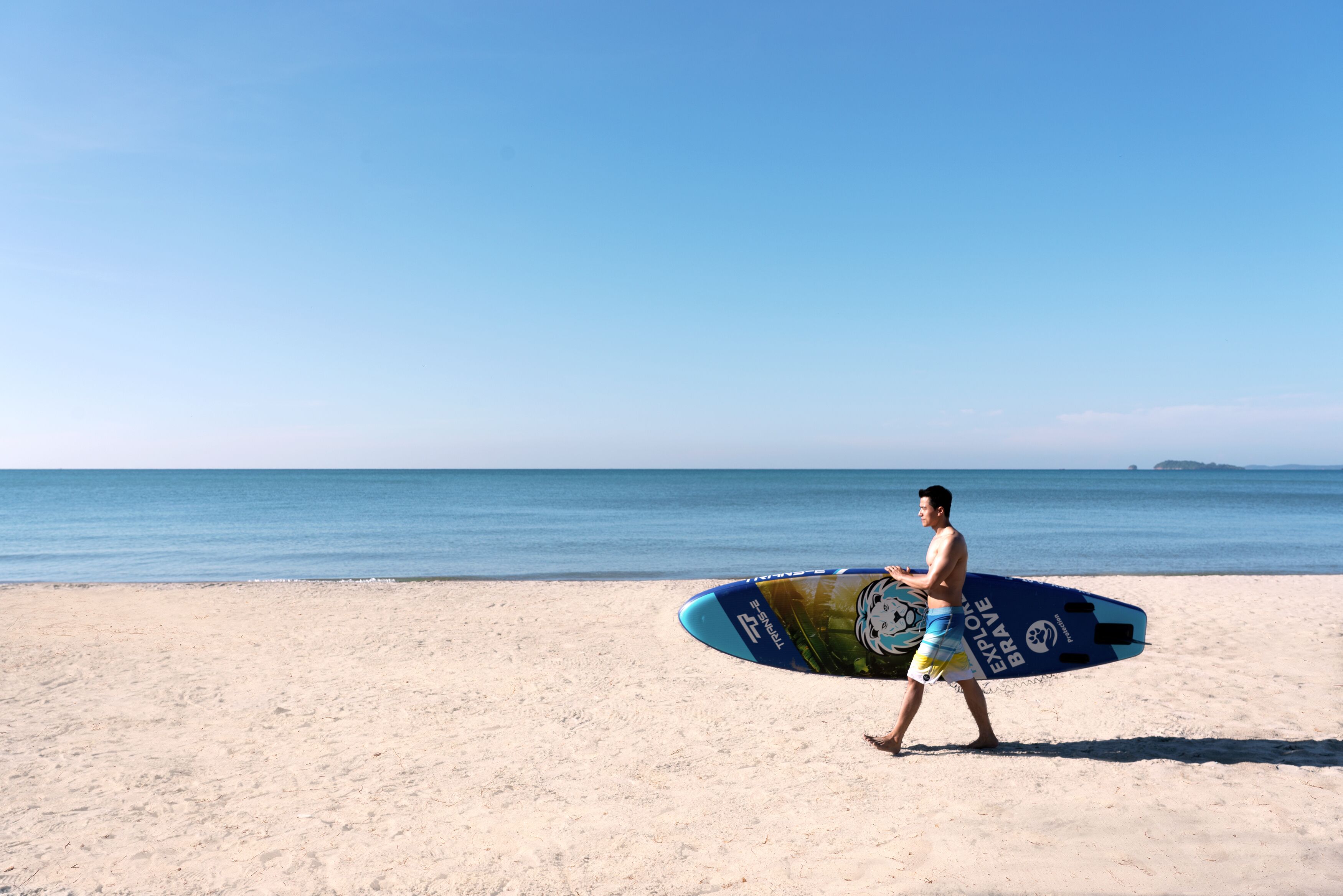 A man carrying a surfboard on a sandy beach with a clear sky and calm sea.