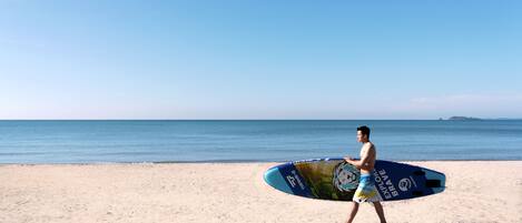 Un hombre lleva una tabla de surf en una playa arenosa, con cielo despejado y mar tranquilo.