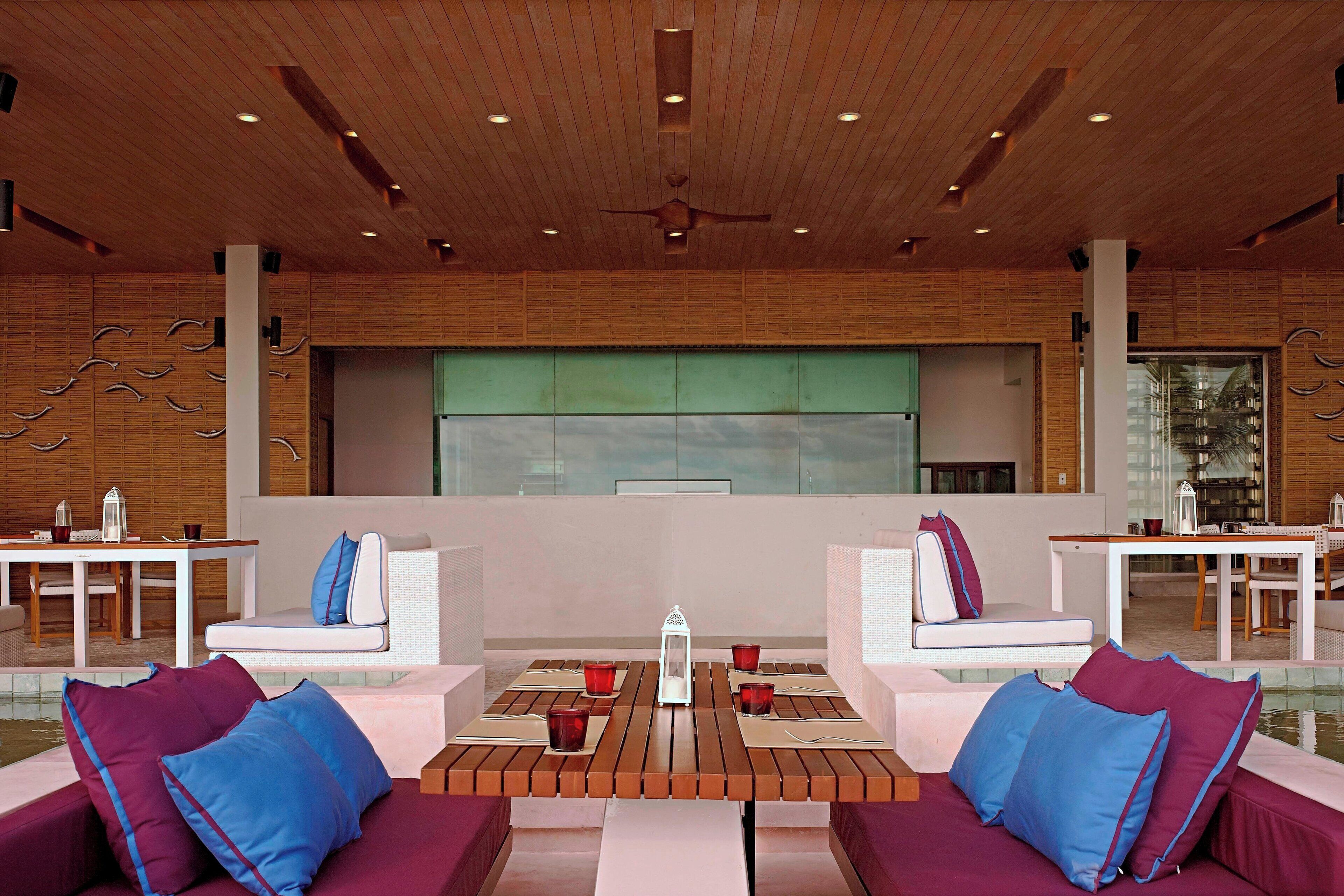 A modern lounge area with wooden ceiling, white sofas with blue cushions, and a central table with red cups.