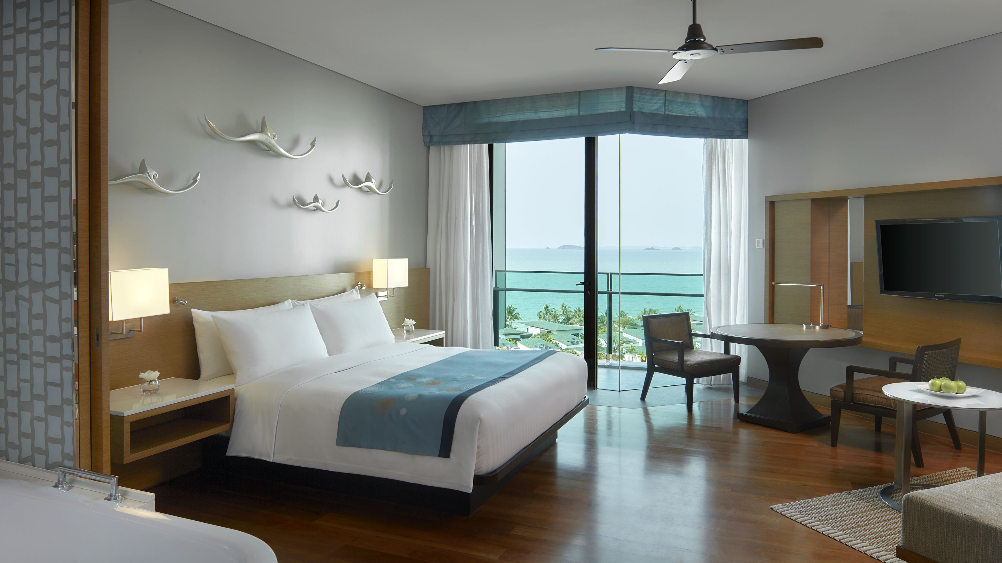 A hotel room with a large bed, a desk with a chair, a TV, and a balcony with a view of the ocean.