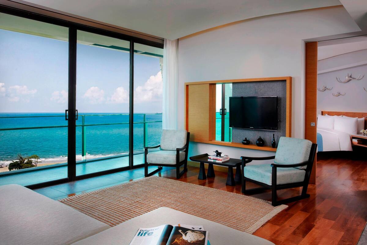 A hotel room with a large sliding glass door leading to a balcony with a view of the ocean. There is a bed, two armchairs, a coffee table, and a flat-screen TV mounted on the wall.