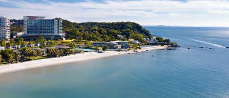A coastal hotel with a beach view, multiple buildings, and a forested hill in the background.