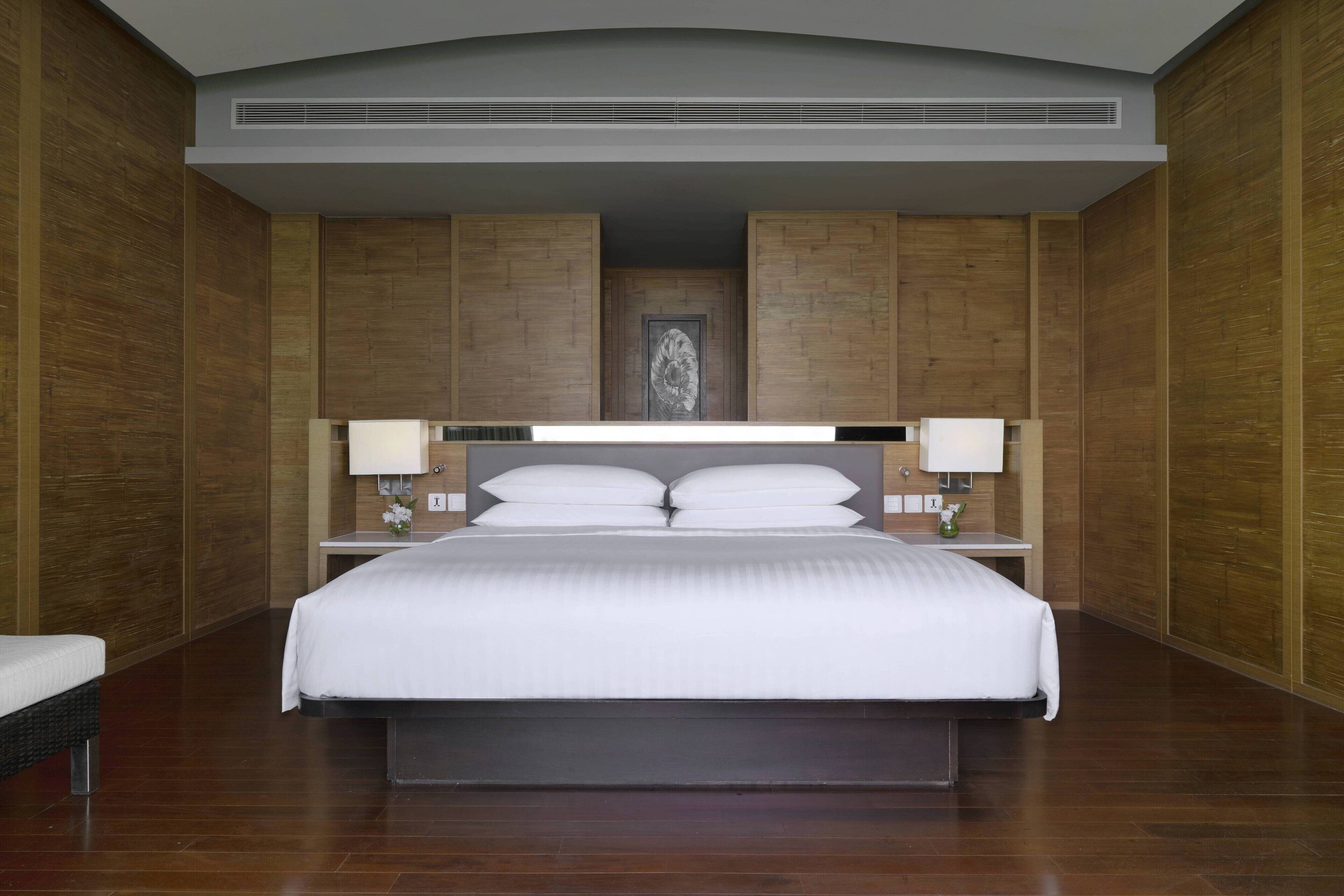 A modern hotel room with a large bed, wooden panel walls, a central artwork, and bedside lamps.