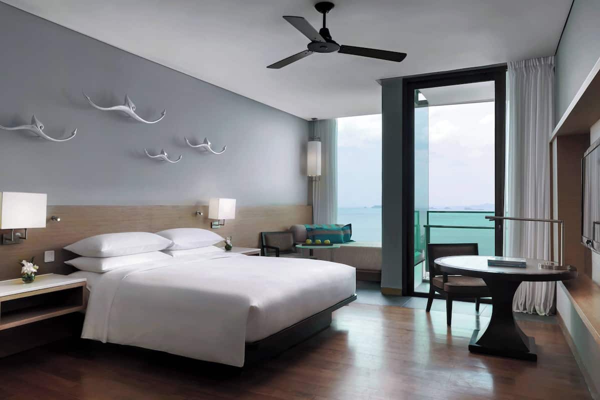 A hotel room with a bed, bedside tables, a desk, a chair, a ceiling fan, and a view of the sea.