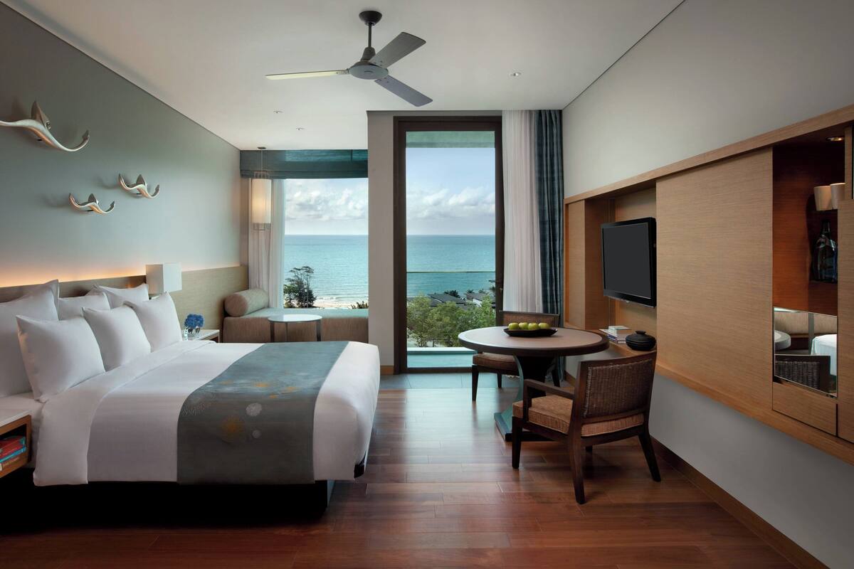 A modern hotel room with a bed, a desk, a chair, a television, and a balcony with a view of the sea.