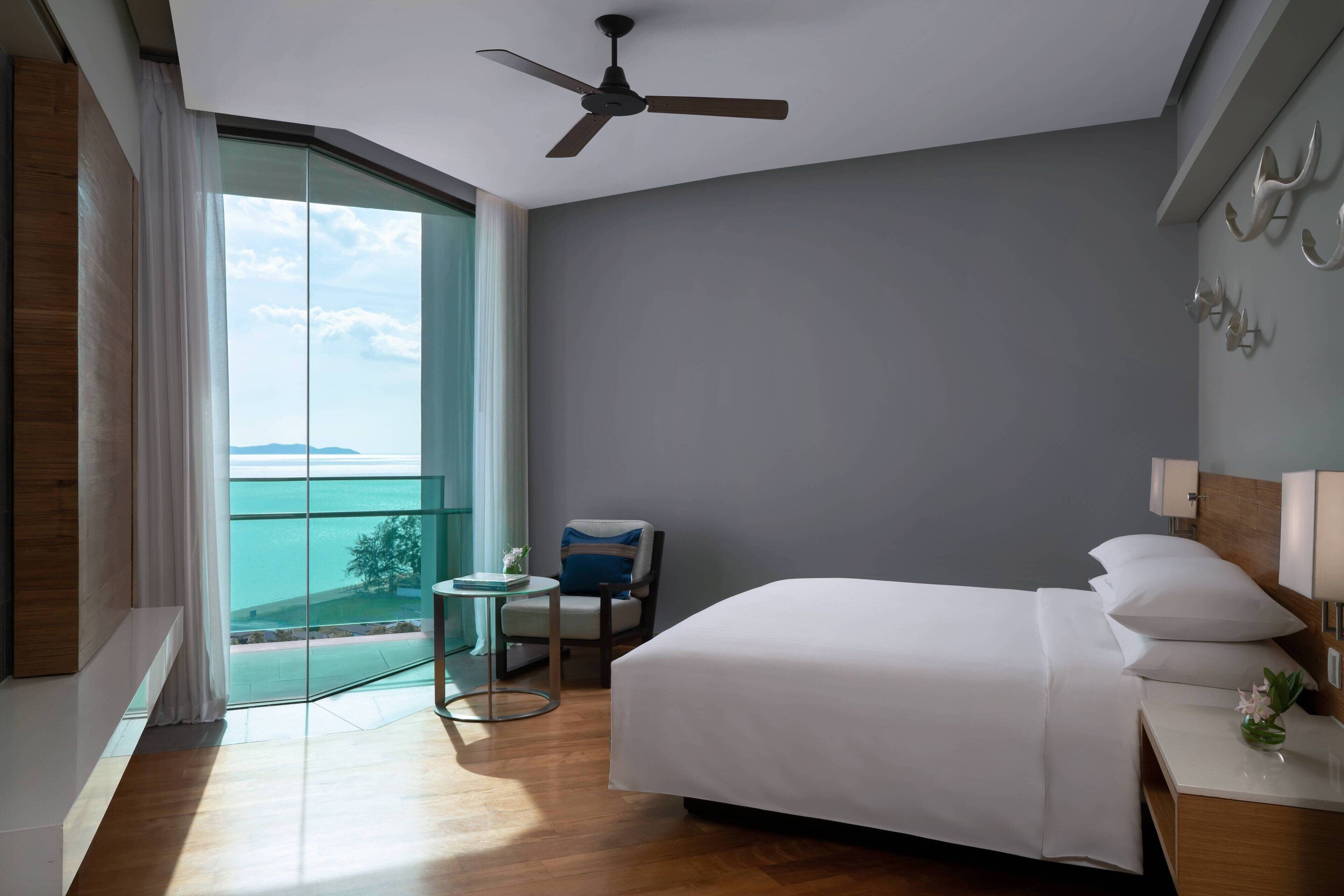 A hotel room with a bed, a small table, a chair, a ceiling fan, and a view of the sea through large windows.