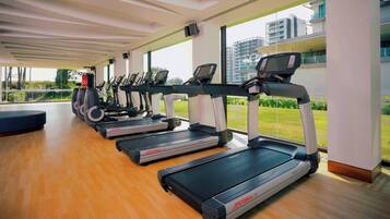 A gym with treadmills aligned along a wall with large windows, a wooden floor, and a view of a building and greenery outside.