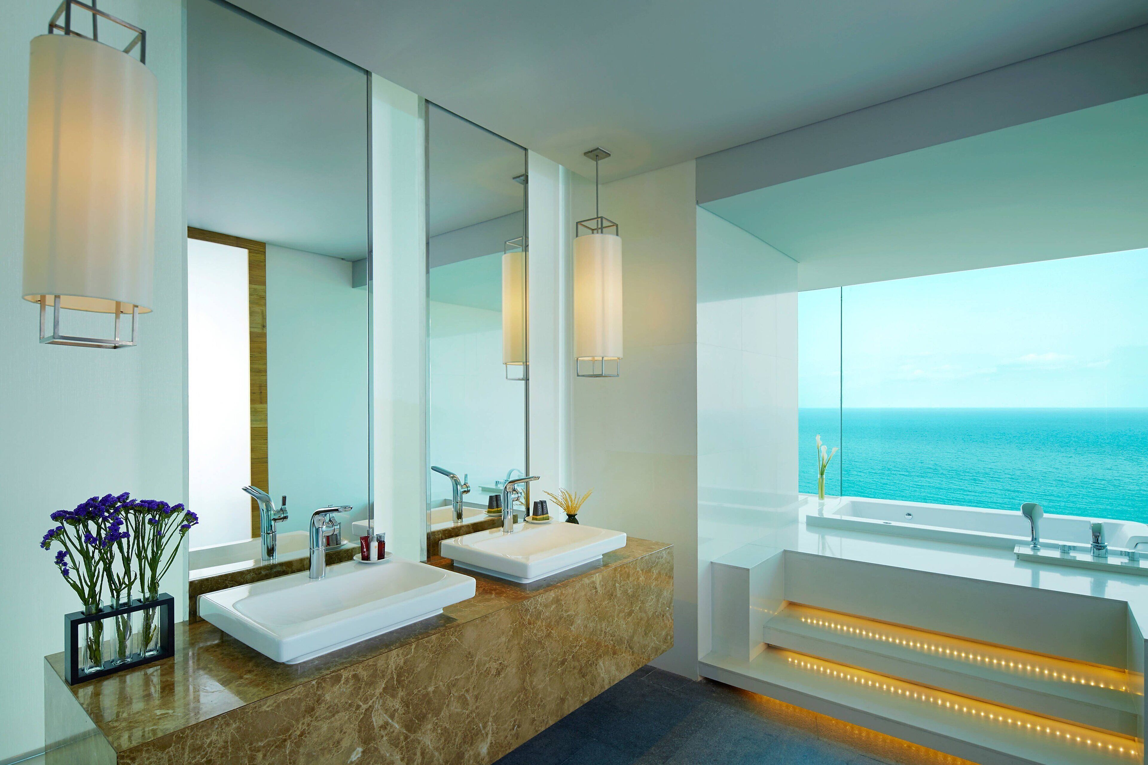 A modern bathroom with two sinks, a large mirror, and a view of the sea.