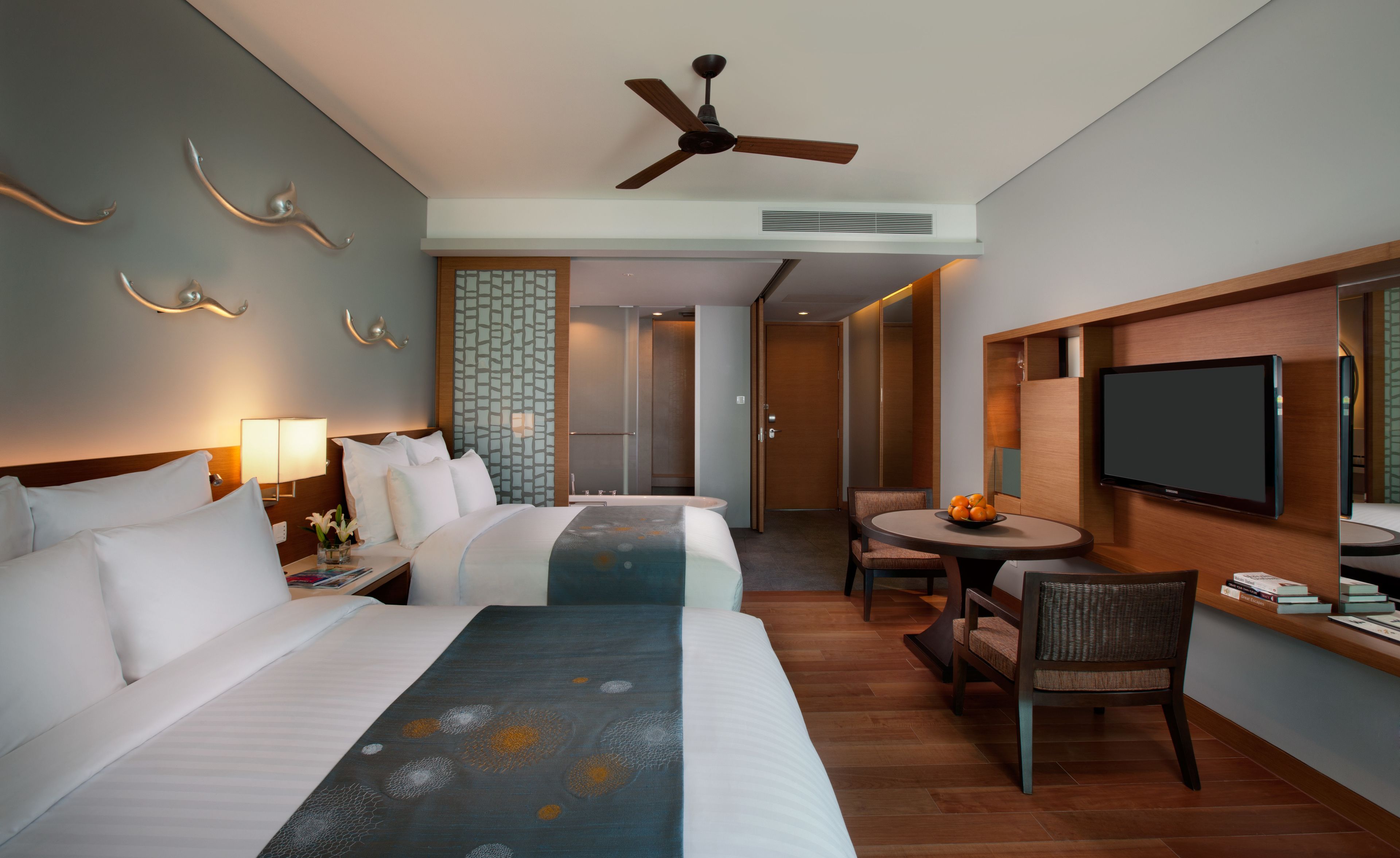 A hotel room with two beds, a ceiling fan, a TV, a small table with fruit, and a chair.
