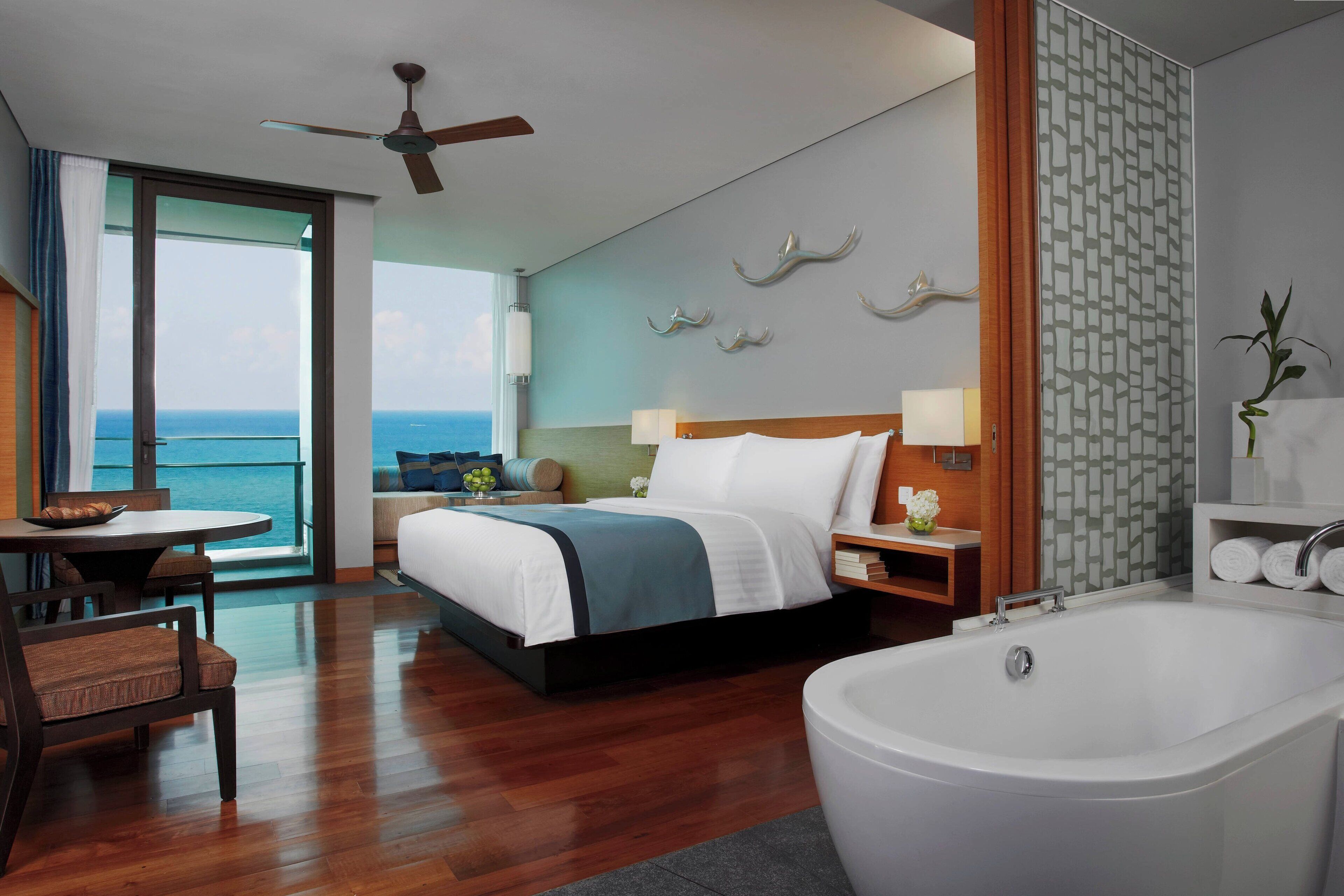 A modern hotel room with a large bed, a sitting area with a view of the sea, a bathroom with a bathtub, and a ceiling fan.