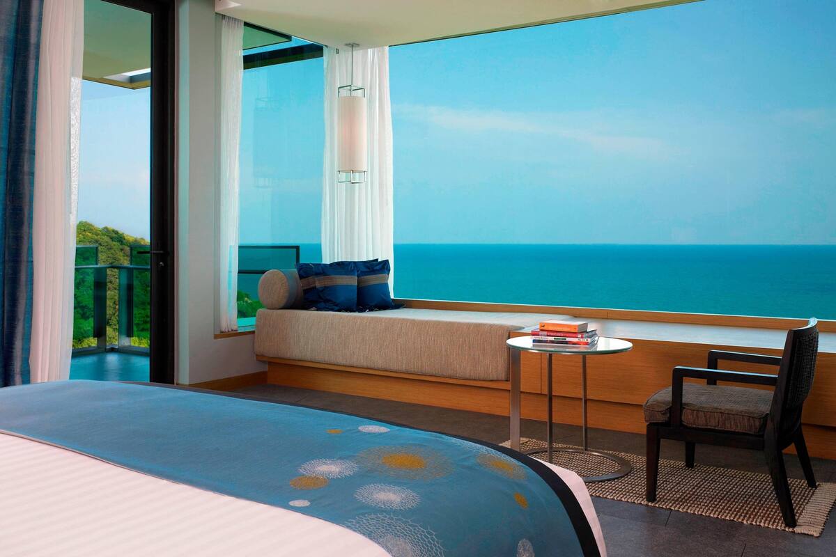 A hotel room with a large bed, a small table with books, a chair, a sofa with a view of the ocean, and a balcony with curtains.