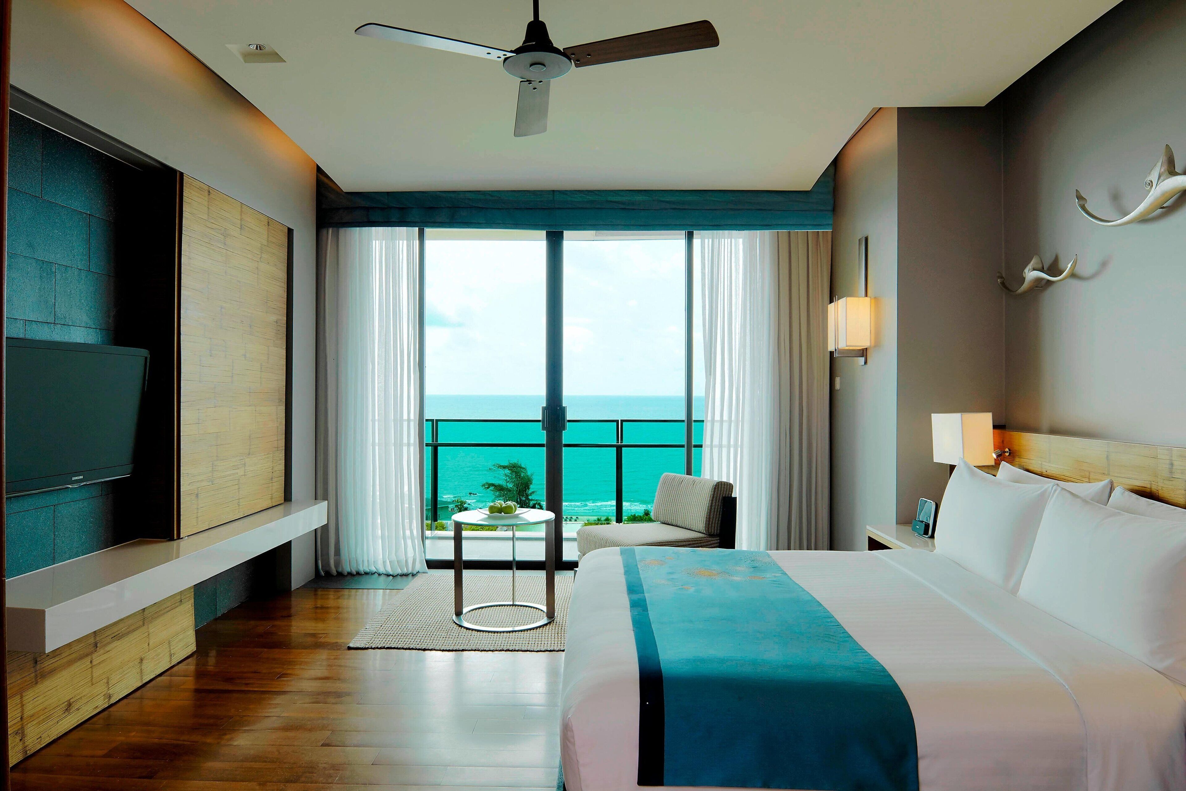 Habitación de hotel con cama, televisión, ventilador de techo, balcón con vista al océano y una mesa con lámpara.