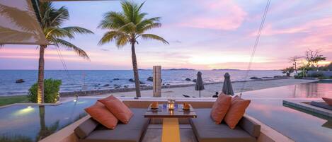 A sunset view from a balcony with a pool, two cushioned seating areas, and a table with a candle.