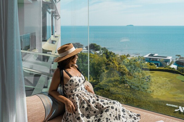 A woman sitting by a large window overlooking a coastal view with a building and greenery.