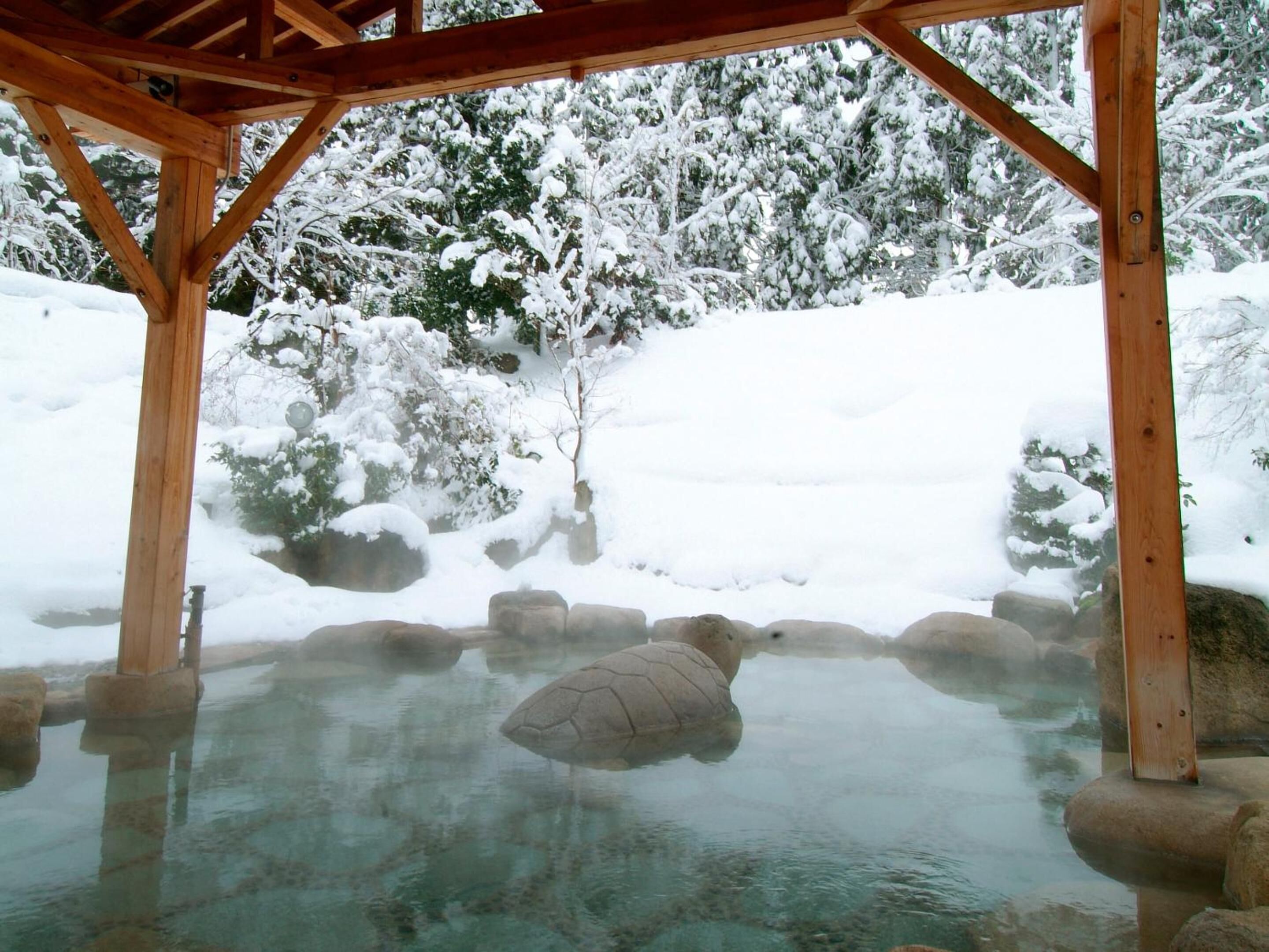 A wooden gazebo with a clear pool surrounded by snow-covered rocks and trees.