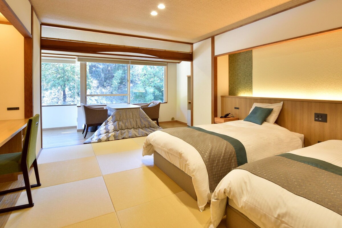 Una habitación de hotel con dos camas, un escritorio y vistas a árboles a través de amplias ventanas.