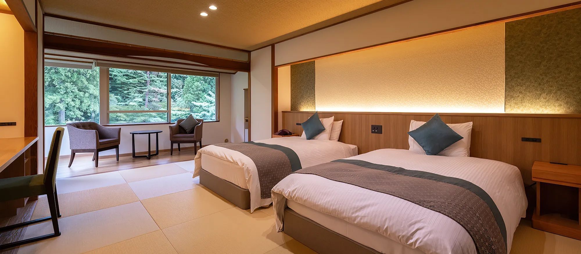 Habitación de hotel con dos camas, un escritorio, una silla y vistas a árboles a través de amplios ventanales.