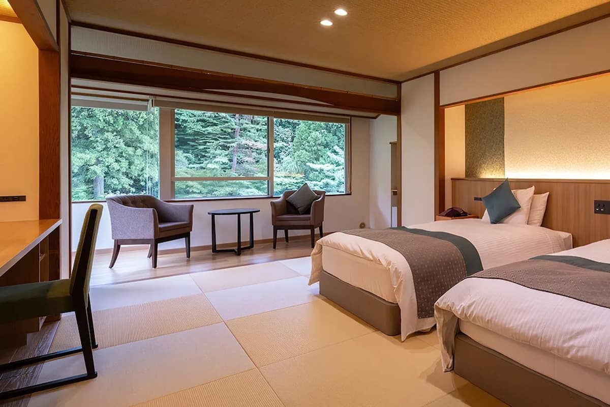 Habitación de hotel con dos camas, un escritorio, una silla y vistas a árboles a través de amplios ventanales.