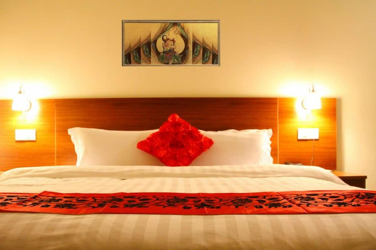 A hotel room with a wooden headboard, two bedside lamps, a large bed with white bedding, a red decorative pillow, and a framed artwork above the bed.