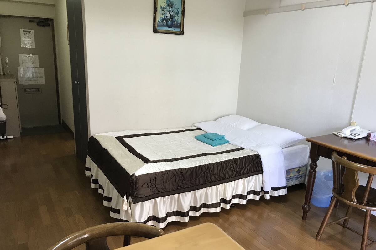 A single bed with a black and white striped bedspread and a teal towel, a wooden table, a chair, and a small desk with a telephone.