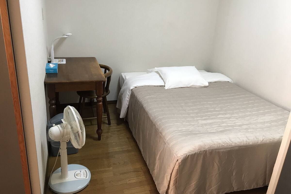 A small, neatly arranged room with a wooden desk, a chair, a bed with white bedding, and a standing fan.