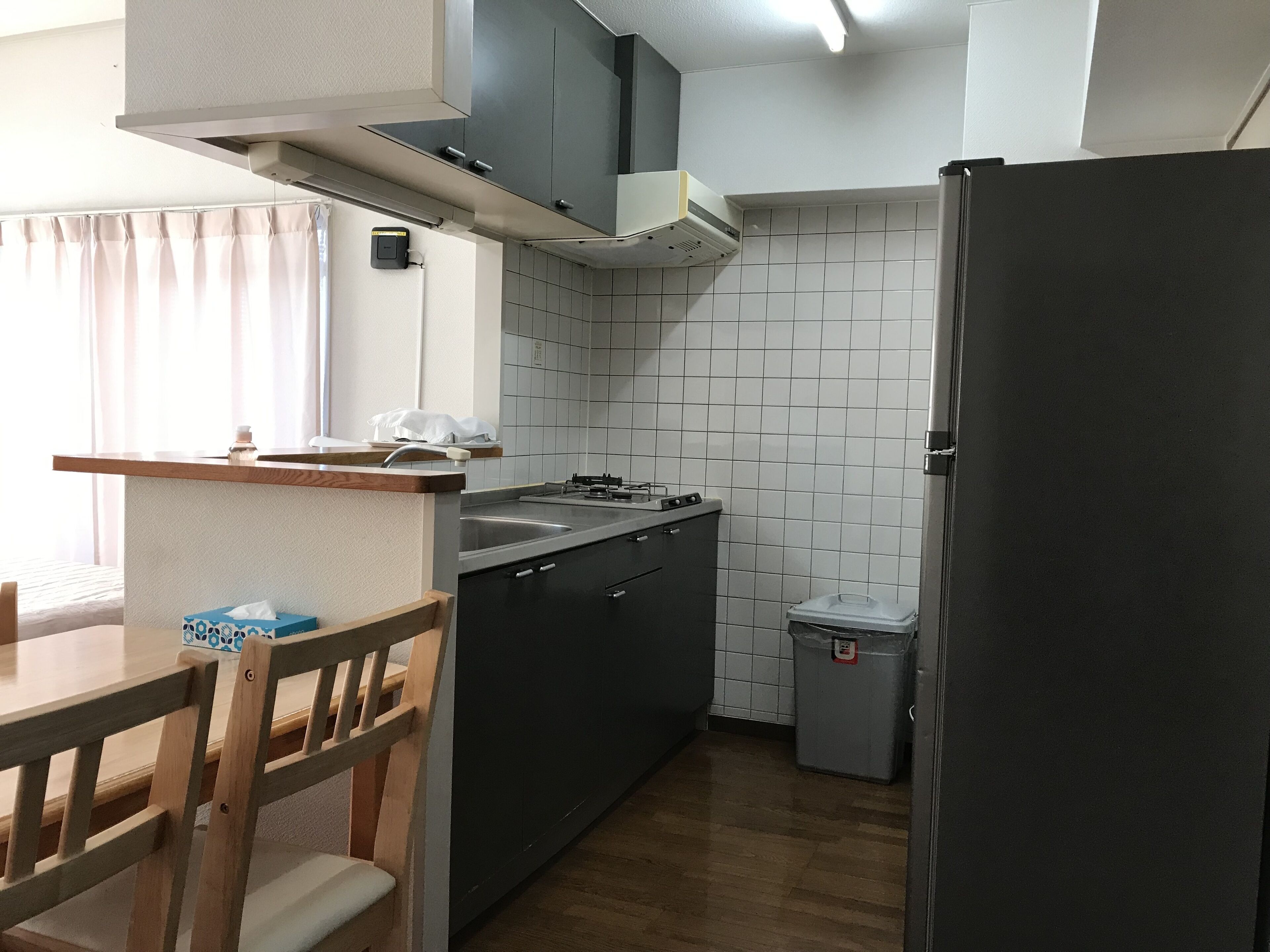 A compact kitchen with a wooden dining table, a refrigerator, a sink, and a trash bin.