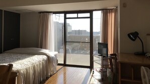 A bedroom with a bed, a desk, a chair, a TV, and a sliding glass door to a balcony. - Weekly Harborview Mansion - Main Building (Naha)