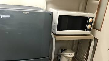 A compact kitchen with a grey refrigerator, a white microwave, and a rice cooker on a shelf.