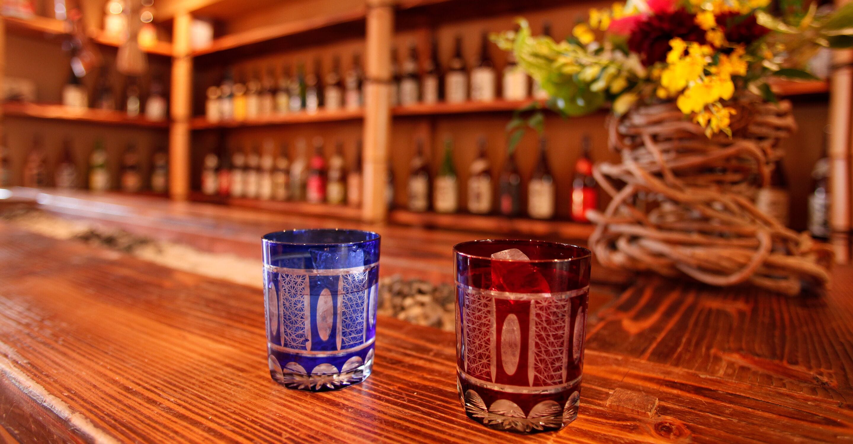 Two glasses on a wooden bar counter with a blurred background of shelves with bottles.