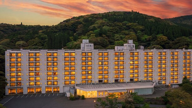 A multi-story hotel with a row of uniformly lit windows, a parking lot, and a hilly backdrop. - Hotel Kirishima Castle (Kirishima)