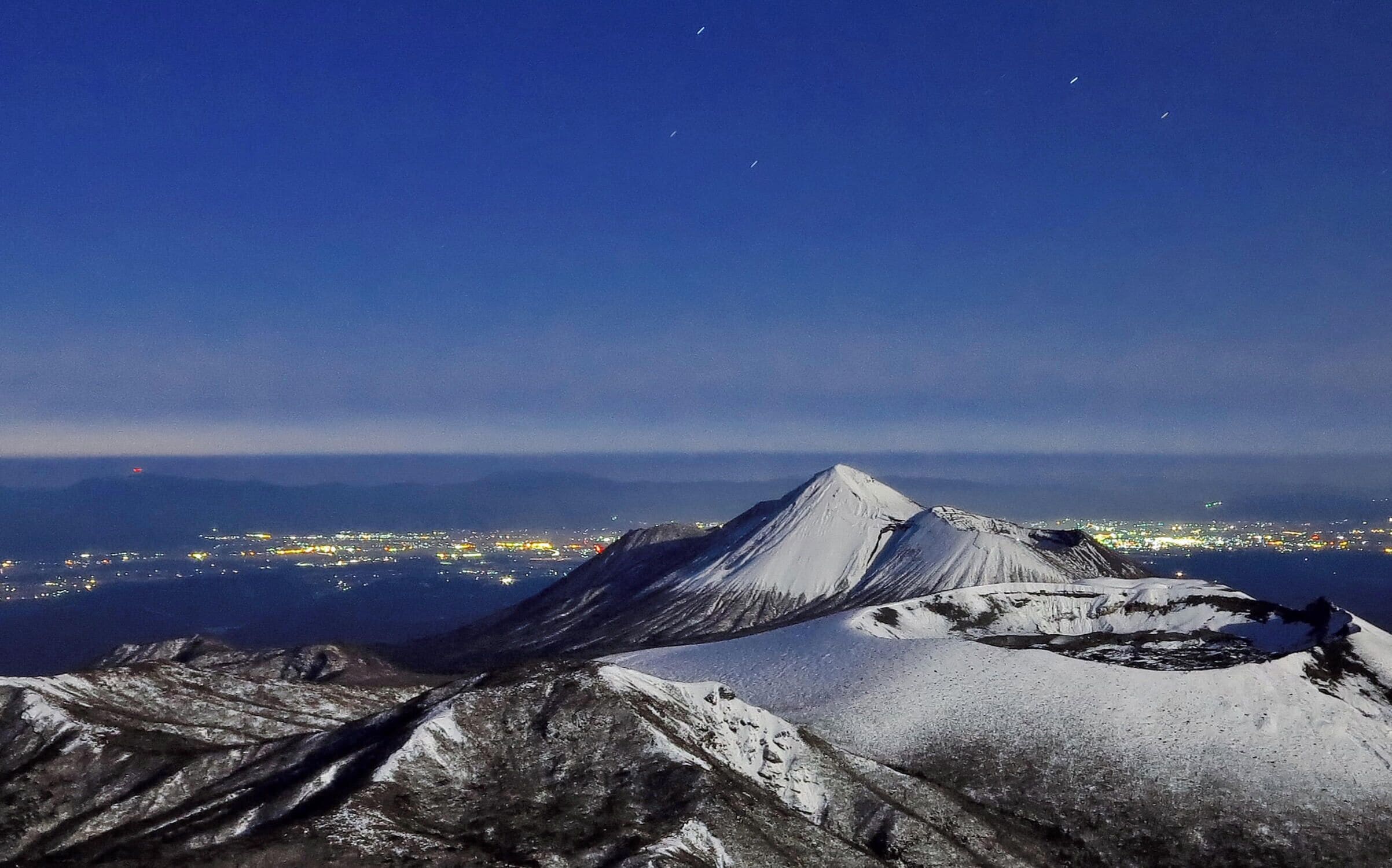 A snow-covered mountain peak at night with city lights in the background.