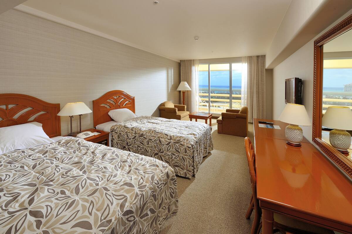 A hotel room with two beds, a desk, a chair, a lamp, and a large window offering a view of the beach.