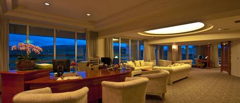 A spacious hotel living room with a large desk, multiple seating areas, and large windows offering a view of the cityscape at dusk.