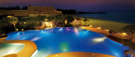 A large, curved swimming pool with lounge chairs and a waterfall feature, surrounded by a landscaped area and a view of the beach at dusk.