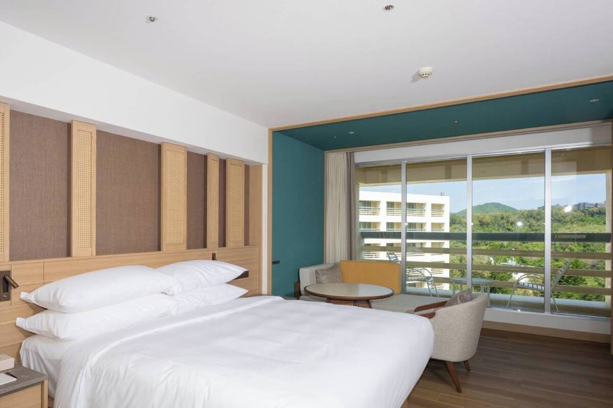 A hotel room with a large bed, a small table, a chair, and a balcony with a view.