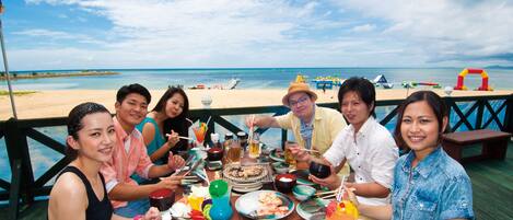 A group of people dining at a wooden deck with a view of the beach and ocean.