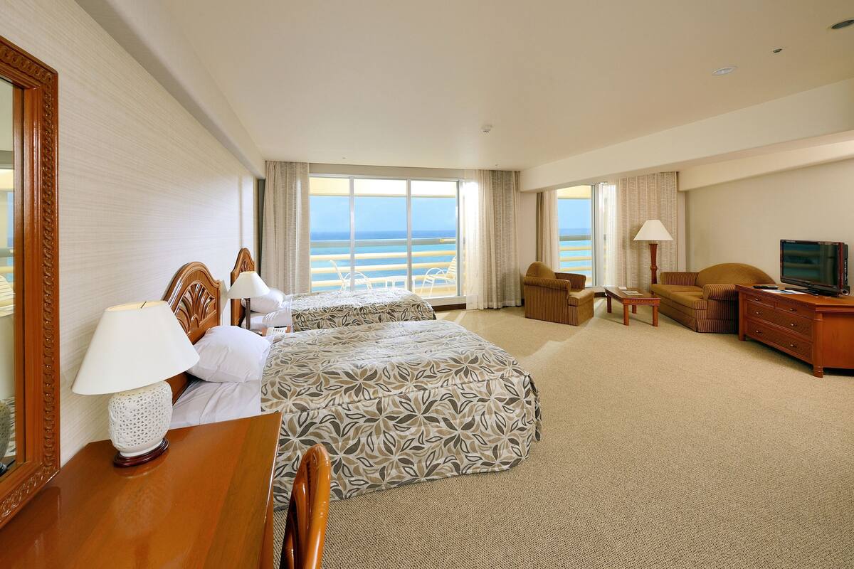A hotel room with a large bed, two bedside tables with lamps, a TV, a sofa, and a view of the beach.