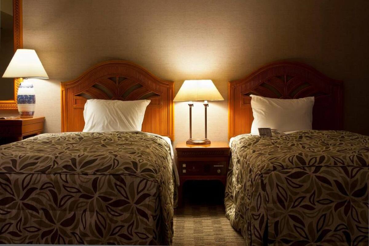 A hotel room with two beds, each with a patterned bedspread and a wooden headboard. There are two bedside lamps and a nightstand with a lamp between the beds.