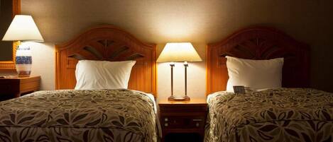 A hotel room with two beds, each with a patterned bedspread and a wooden headboard. There are two bedside lamps and a nightstand with a lamp between the beds.