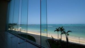 A view from a window overlooking a beach with clear blue water, palm trees, and a sandy shore. - Rizzan Sea Park Hotel Tancha Bay (Onna)