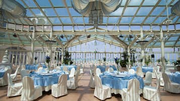 A large, open-air venue with tables set for events, featuring a glass ceiling and a view of the beach.