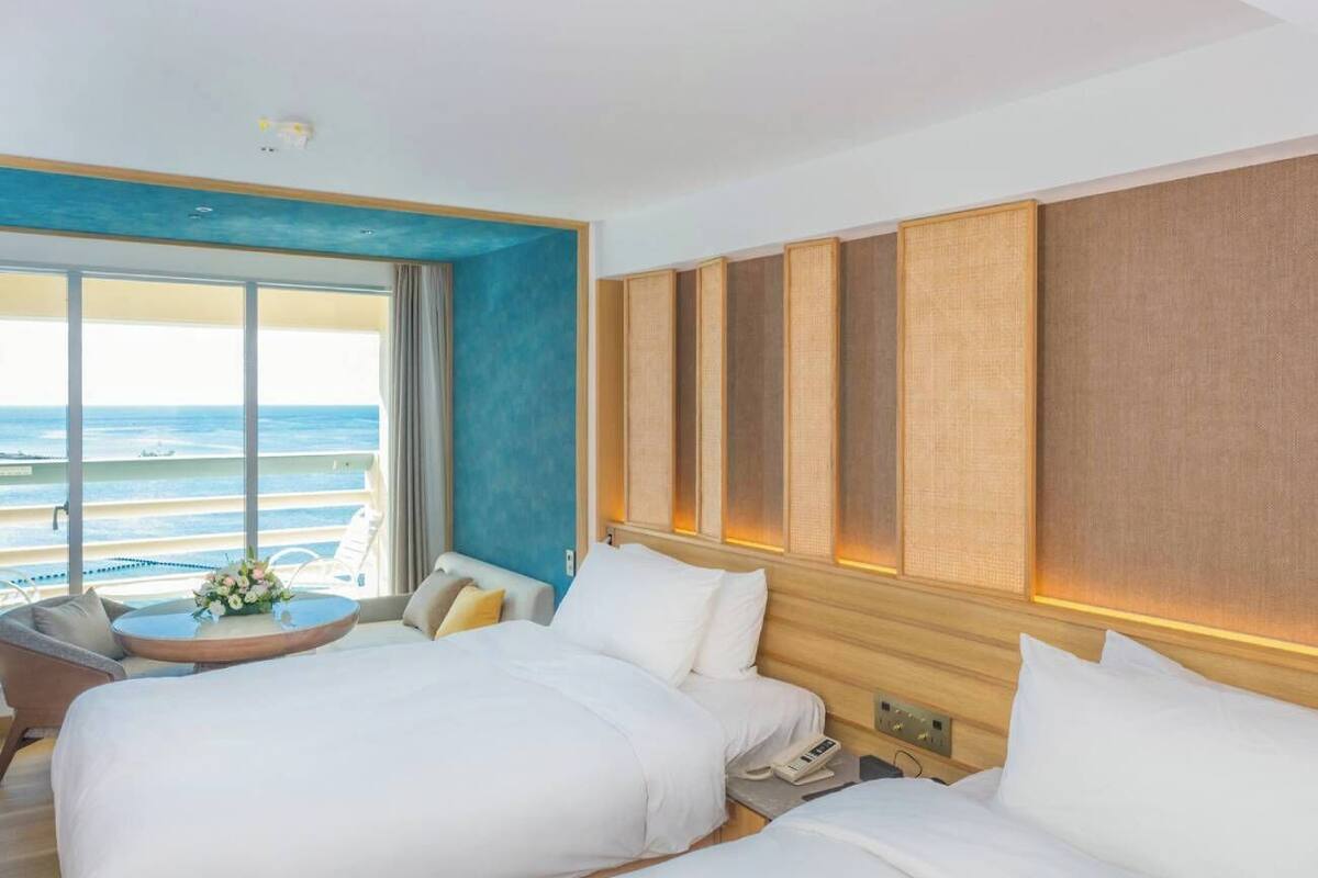 A hotel room with two beds, a view of the ocean, a small table with flowers, and a seating area with a chair.