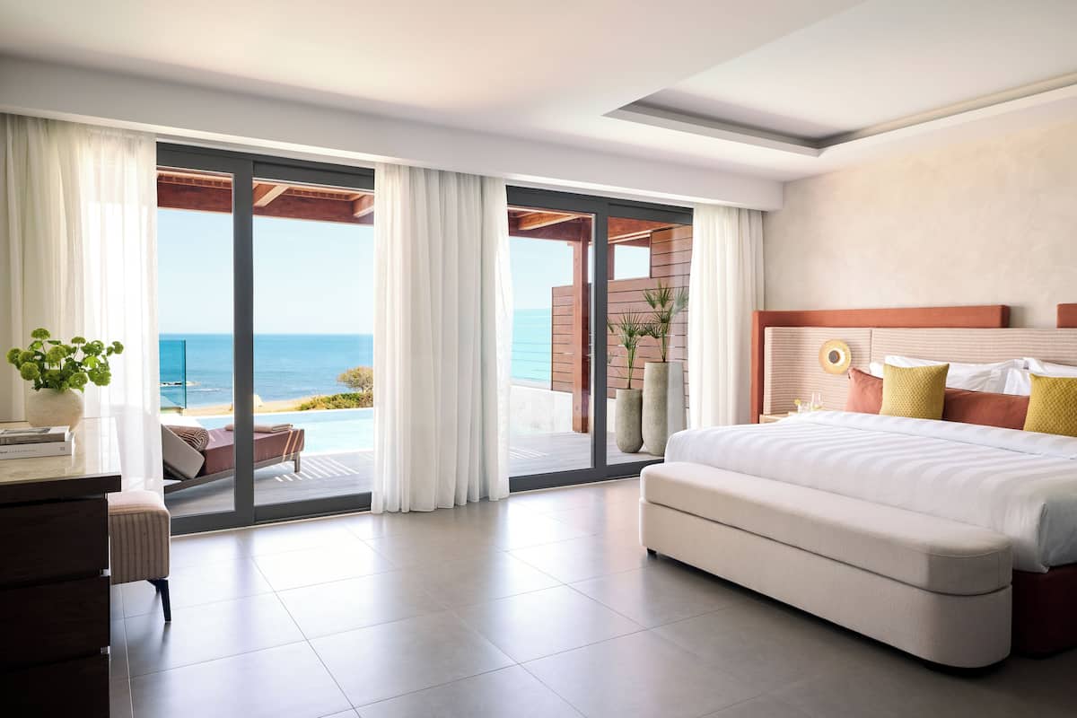 Ein modernes Hotelzimmer mit einem großen Bett, einem Schreibtisch und Meerblick durch raumhohe Fenster.