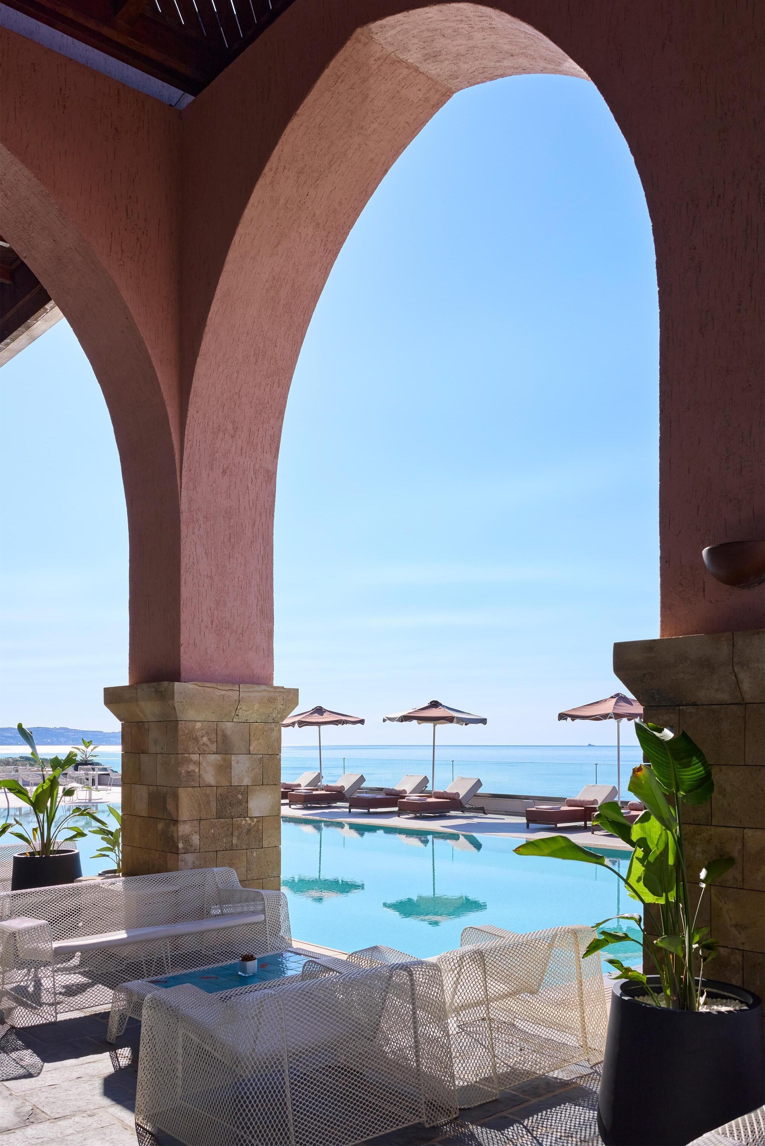A poolside area with lounge chairs and umbrellas, overlooking a clear blue sea.